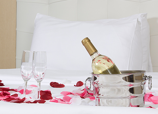 wine bottle sitting in a metal ice bucket on a bed with two glasses and rose petals on bed