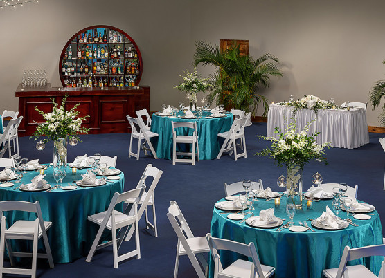 indoor event space area with blue tablecloth round tables, white folding chairs, and flower table centerpieces
