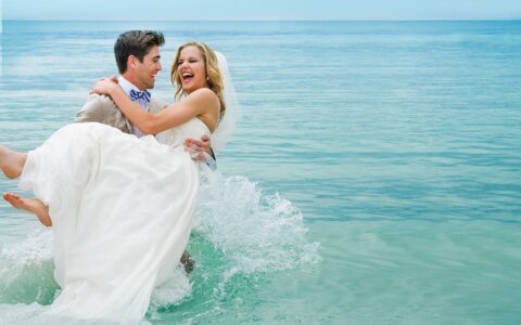 groom holding the bride as he walks through the ocean water