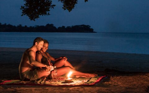 couple sitting on a blanket with a candle having drinks on the beach at night